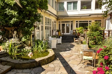 Oshos Place Ein ruhiger Innenhof mit einem kleinen, von Pflanzen umgebenen Teich. Sitzmöglichkeiten im Freien laden zum Verweilen ein, während üppiges Grün den Charme des Ortes unterstreicht.