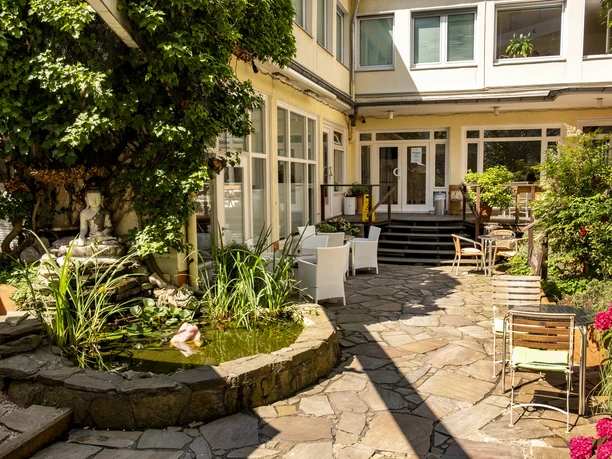 Oshos Place Ein ruhiger Innenhof mit einem kleinen, von Pflanzen umgebenen Teich. Sitzmöglichkeiten im Freien laden zum Verweilen ein, während üppiges Grün den Charme des Ortes unterstreicht.