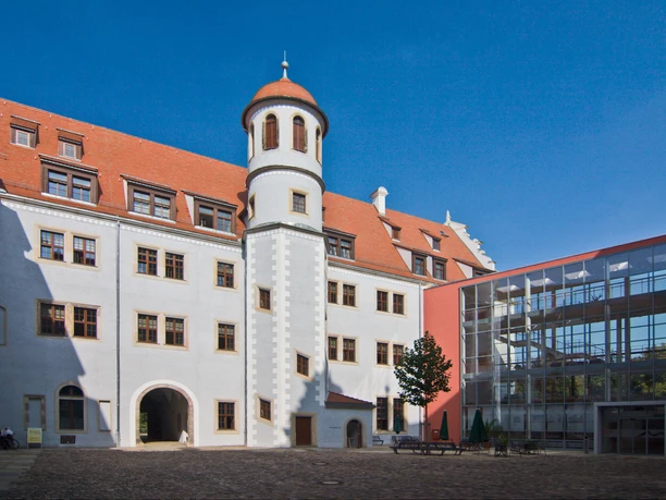 zwickau-osterstein-06-c-mattrose-de.jpg Historisches Gebäude mit Turm im Zentrum eines gepflasterten Hofs, daneben moderne Glasstruktur.