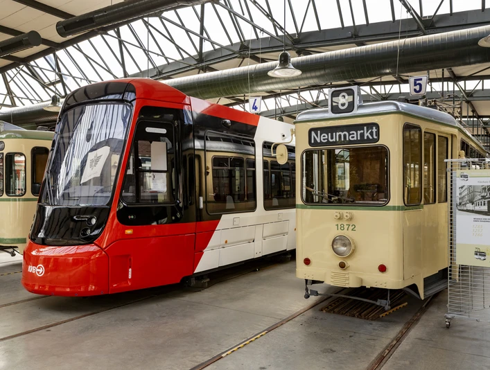 Thielenbruch Tram Museum Three historic streetcars stand side by side in a hall: two older models with cream and green paintwork and a modern red streetcar. Signs above the vehicles indicate stops such as "Neumarkt". The light-filled hall is made of steel and glass.