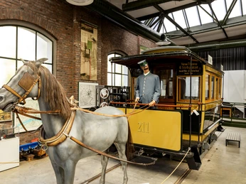Strassenbahn-Museum-Thielenbruch In einem historischen Straßenbahnmuseum steht eine alte Pferdebahn mit realistisch dargestelltem Pferd und Kutscher. Die Szene wird von einer alten Backsteinhalle umgeben, die historische Plakate und Exponate beherbergt, was den Charme der vergangenen Transportzeiten eindrucksvoll einfängt.