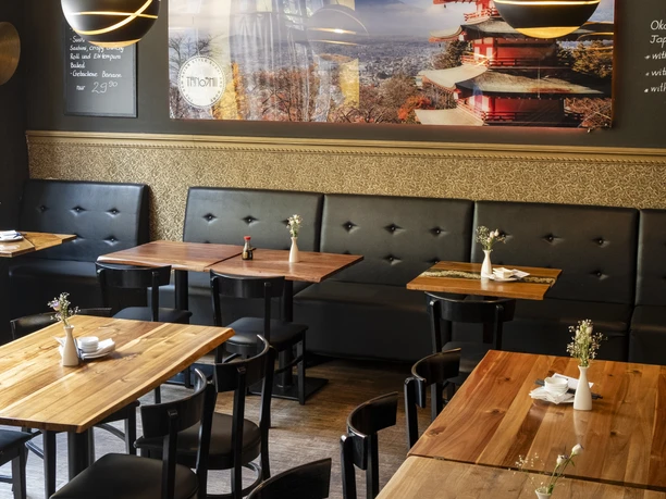 Ein stilvoll eingerichtetes Restaurant mit hölzernen Tischen, modernen Lampen und einem beeindruckenden Wandbild eines japanischen Tempels im Herbst.