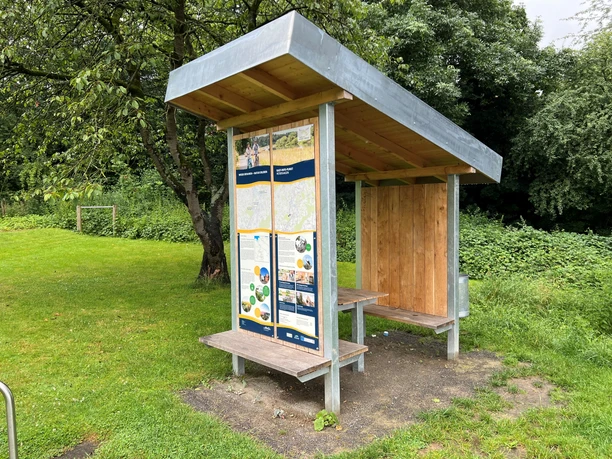 Rastplatz Petershagen Wetterschutz mit Bänken an einer Wiese, ergänzt durch Informationstafeln unter einem Schrägdach.