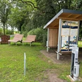 Rastplatz Hopfenberg Rastplatz mit überdachten Infotafeln, zwei Liegebänken und einer Ladesäule, umgeben von Bäumen.