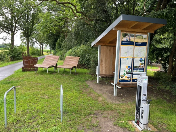 Rastplatz Hopfenberg Rastplatz mit überdachten Infotafeln, zwei Liegebänken und einer Ladesäule, umgeben von Bäumen.