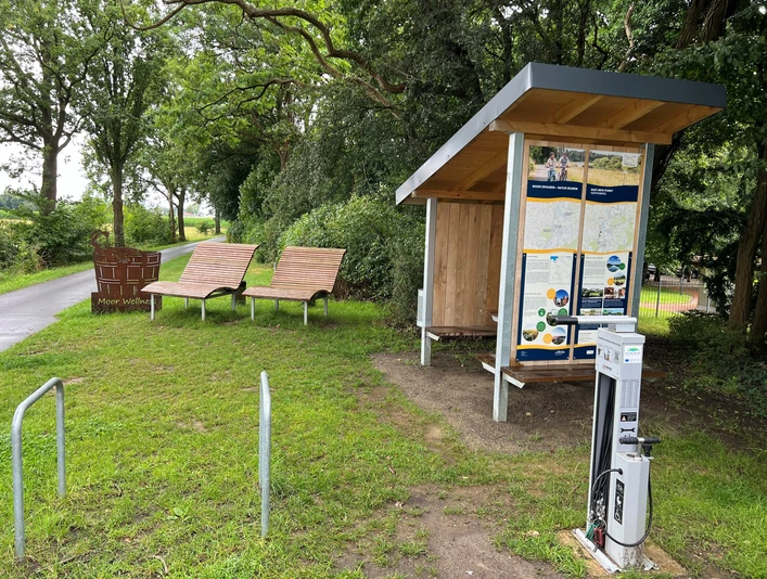 Rastplatz Hopfenberg Rastplatz mit überdachten Infotafeln, zwei Liegebänken und einer Ladesäule, umgeben von Bäumen.