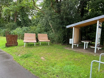 Rastplatz Hopfenberg Zwei Holzliegen und ein Unterstand mit Tisch im Grünen, umgeben von Bäumen und einem Weg.