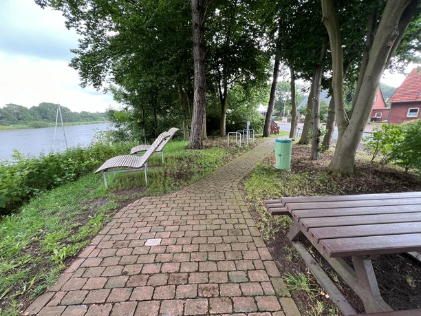 Rastplatz Heimsen Ein schmaler Pflasterweg führt zu Holzliegen und einem Tisch inmitten von Bäumen am Flussufer.