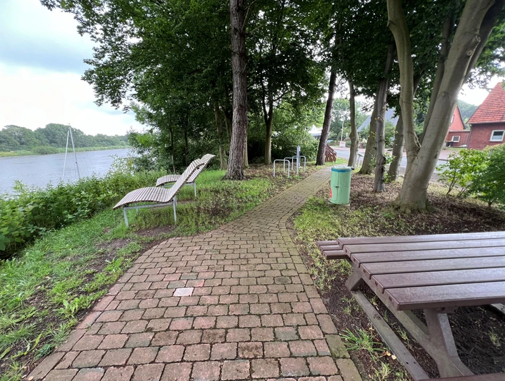 Rastplatz Heimsen Ein schmaler Pflasterweg führt zu Holzliegen und einem Tisch inmitten von Bäumen am Flussufer.