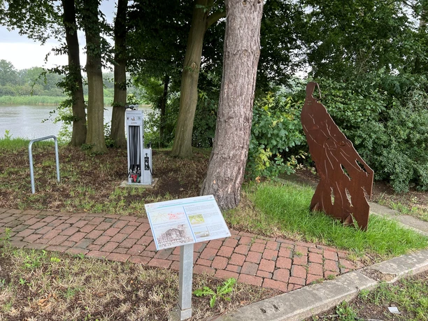 Rastplatz Heimsen Rastplatz am Waldrand mit Skulptur und Infotafel; im Hintergrund fließt ein ruhiger Fluss vorbei.