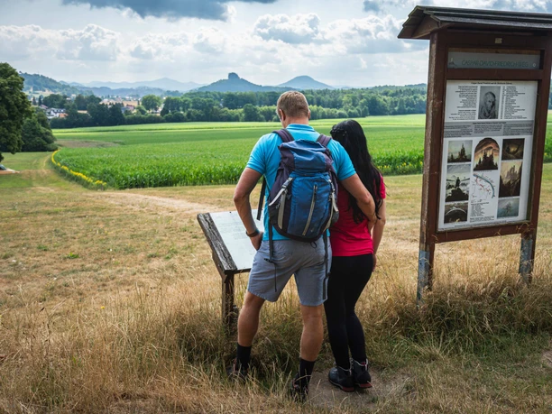 Caspar-David-Friedrich-Weg am Wolfsberg Ein Paar steht vor einer Informationstafel auf einem Wanderweg mit Blick auf grüne Felder und bewaldete Hügel unter bewölktem Himmel.