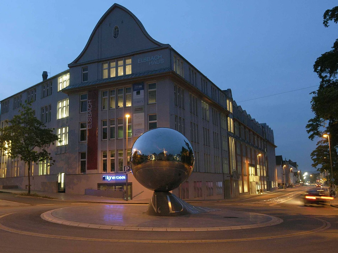 Eine glänzende, silberne Kugel auf einem kreisförmigen Sockel steht vor einem beleuchteten Gebäude bei Nacht.