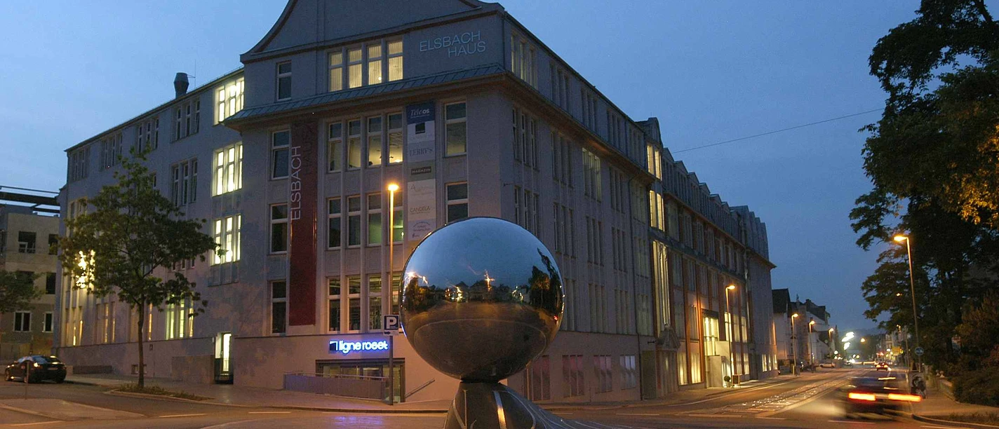 Eine glänzende, silberne Kugel auf einem kreisförmigen Sockel steht vor einem beleuchteten Gebäude bei Nacht.