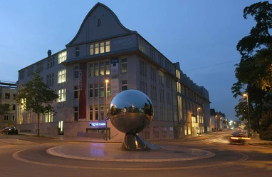 Kugel_Elsbach_quer_Nacht.jpg Eine glänzende, silberne Kugel auf einem kreisförmigen Sockel steht vor einem beleuchteten Gebäude bei Nacht.