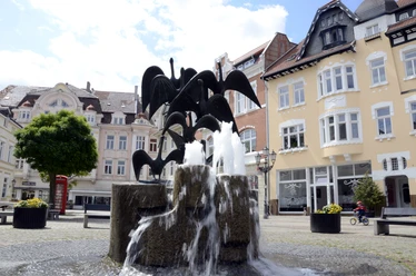 Gänsemarkt_2011_Siwinski_4.jpg Ein Brunnen mit Gänsestatuen vor historischen Gebäuden bei sonnigem Wetter auf einem Platz.