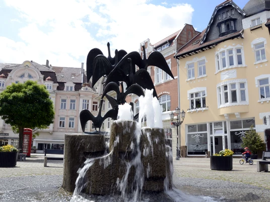 Gänsemarkt_2011_Siwinski_4.jpg Ein Brunnen mit Gänsestatuen vor historischen Gebäuden bei sonnigem Wetter auf einem Platz.