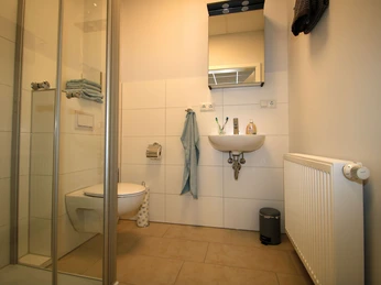 FeWo-thelivingroom-dusche-wc.jpg Badezimmer mit Dusche, WC, Waschbecken und Handtuchhalter. Helles Design mit neutralen Fliesen.