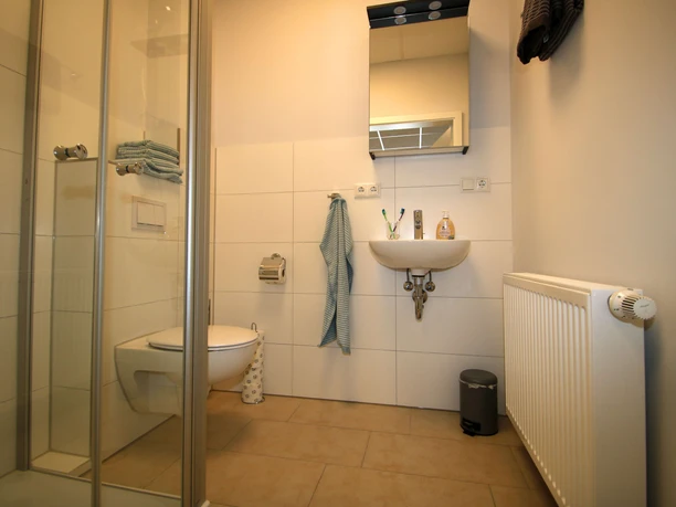 FeWo-thelivingroom-dusche-wc.jpg Badezimmer mit Dusche, WC, Waschbecken und Handtuchhalter. Helles Design mit neutralen Fliesen.