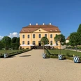 Blick zum Schloss Gelbes Barockgebäude des Barockgarten Großsedlitz mit rotem Dach, umgeben von gepflegtem Garten und Bäumen in blau-weißen Töpfen, unter klarem blauem Himmel.