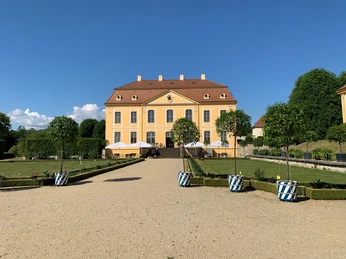Blick zum Schloss Gelbes Barockgebäude des Barockgarten Großsedlitz mit rotem Dach, umgeben von gepflegtem Garten und Bäumen in blau-weißen Töpfen, unter klarem blauem Himmel.Yellow baroque building of the Grosssedlitz Baroque Garden with a red roof, surrounded by well-tended gardens and trees in blue and white pots, under a clear blue sky.Žlutá barokní budova barokní zahrady Grosssedlitz s červenou střechou, obklopená udržovanými zahradami a stromy v modrých a bílých květináčích, pod jasně modrou oblohou.Żółty barokowy budynek Ogrodu Barokowego Grosssedlitz z czerwonym dachem, otoczony zadbanymi ogrodami i drzewami w niebieskich i białych donicach, pod czystym błękitnym niebem.Geel barokgebouw van de Grosssedlitz Baroque Garden met een rood dak, omringd door goed onderhouden tuinen en bomen in blauwe en witte potten, onder een strakblauwe hemel.Edificio barocco giallo del Giardino Barocco di Grosssedlitz con tetto rosso, circondato da giardini ben curati e alberi in vasi blu e bianchi, sotto un cielo azzurro e limpido.