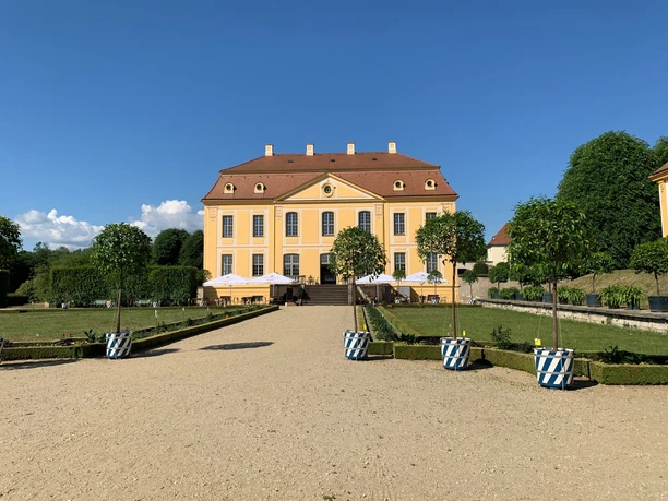 Blick zum Schloss Gelbes Barockgebäude des Barockgarten Großsedlitz mit rotem Dach, umgeben von gepflegtem Garten und Bäumen in blau-weißen Töpfen, unter klarem blauem Himmel.