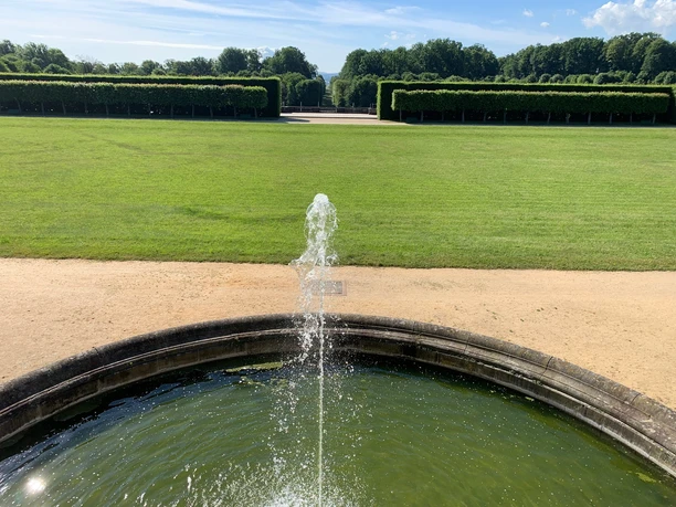 Park Baroque Garden Großsedlitz Fountain in the Grosssedlitz Baroque Garden with a green meadow and clipped hedges in the background under a blue sky.
