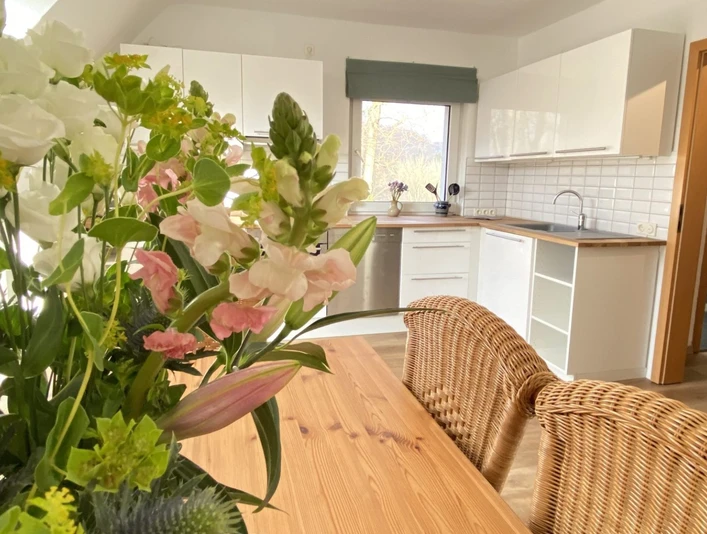 Die Küche in der Fewo-Neandersteig in Heiligenhaus Helle Küche in Heiligenhaus mit Esstisch, Korbstühlen, Blumenarrangement und moderner Ausstattung.