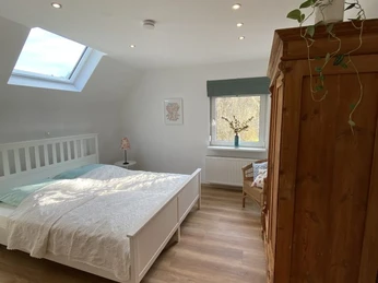 Schlafzimmer in der Fewo-Neandersteig in Heiligenhaus Helles Schlafzimmer mit Holzfußboden, Doppelbett, Kleiderschrank und seitlichem Fenster unter der Dachschräge.