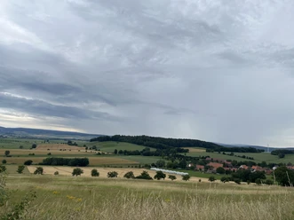 Panoramaweg, Wanderweg, Aussicht Avendshausen ©Einbeck Tourismus.jpeg Panoramablick über weite Felder auf das Dorf Avendshausen unter bewölktem Himmel.