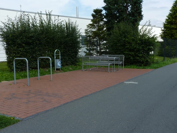 Ruhiger Rastplatz mit Metallbank und Fahrradständern auf einer gepflasterten Fläche neben einem Weg.