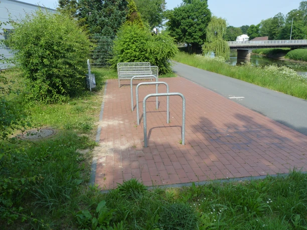 Fahrradständer auf einem gepflasterten Platz, umgeben von grünen Sträuchern und einem ruhigen Kanal.