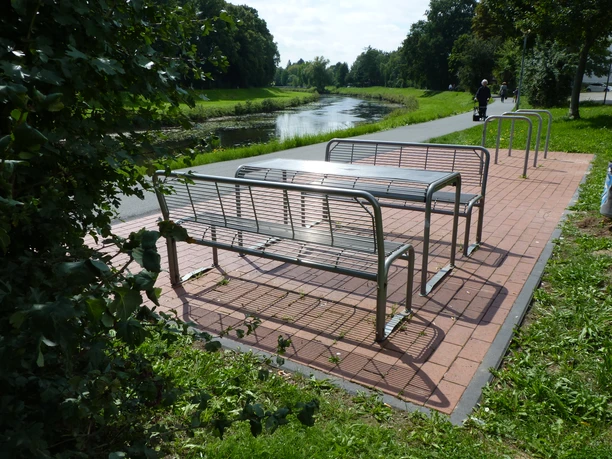 Drei moderne Metallbänke auf gepflastertem Bereich an einem ruhigen Flussufer mit schattenspendenden Bäumen.