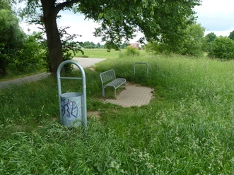 P1110029.JPG Eine einsame Parkbank auf einer kleinen gepflasterten Fläche, umgeben von hohem Gras und einem Plastikmülleimer.
