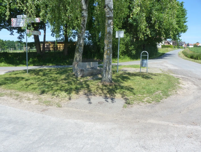 Kreuzung ländlicher Straßen mit Bank unter Bäumen, umgeben von Feldern und einem Abfalleimer.