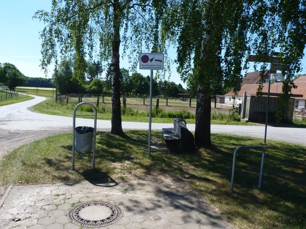 Ein ruhiger Rastplatz mit einer Holzbank unter Bäumen neben einer Landstraße an einem sonnigen Tag.