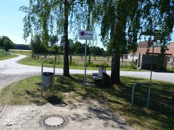 P1110099.JPG Ein ruhiger Rastplatz mit einer Holzbank unter Bäumen neben einer Landstraße an einem sonnigen Tag.