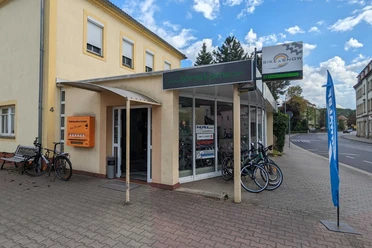 Bike & Snow Fahrradverleih Pirna Fahrradgeschäft mit gelber Fassade, Schaufenster voller Fahrräder, orangem Schlauchautomaten und blauer Flagge auf Gehweg bei bewölktem Himmel.