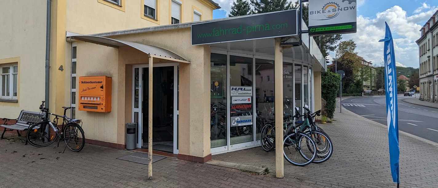 Bike & Snow Fahrradverleih Pirna Fahrradgeschäft mit gelber Fassade, Schaufenster voller Fahrräder, orangem Schlauchautomaten und blauer Flagge auf Gehweg bei bewölktem Himmel.