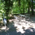 Drei Metallbänke mit Tisch stehen im Schatten grüner Bäume, daneben ein Mülleimer auf Waldboden.