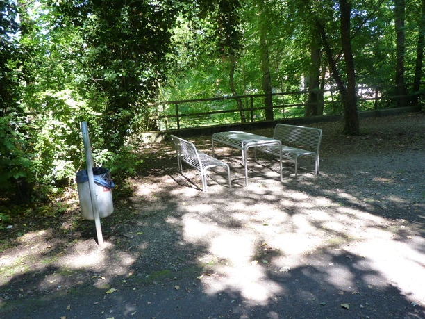 Drei Metallbänke mit Tisch stehen im Schatten grüner Bäume, daneben ein Mülleimer auf Waldboden.