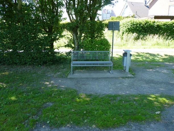 Eine einfache Metallbank steht auf einem verlassenen Weg, umgeben von hoher Hecke und grünem Gras.