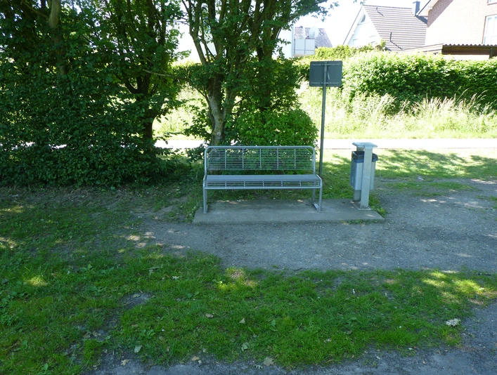 Eine einfache Metallbank steht auf einem verlassenen Weg, umgeben von hoher Hecke und grünem Gras.