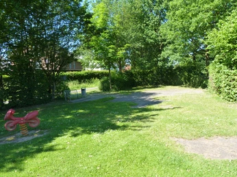 Grüne Parkanlage mit Spielzeugwippe für Kinder, umgeben von Bäumen und einer Sitzbank im Schatten.