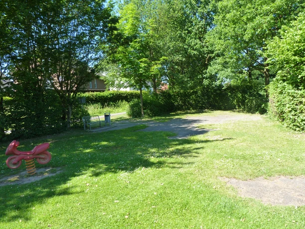 Grüne Parkanlage mit Spielzeugwippe für Kinder, umgeben von Bäumen und einer Sitzbank im Schatten.