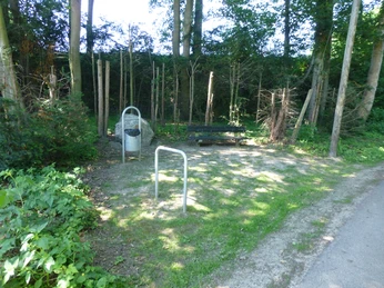 Grüne Parkbank im Schatten, umgeben von Bäumen, daneben ein Abfalleimer und ein Fahrradständer.