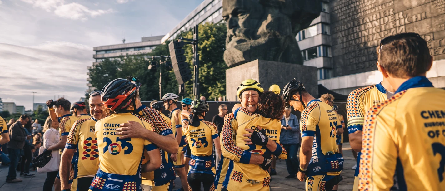 european-peace-ride-opener-ernesto-uhlmann.jpg Eine Gruppe Radfahrer in gelben Trikots steht vor einem großen steinernen Kopfdenkmal.
