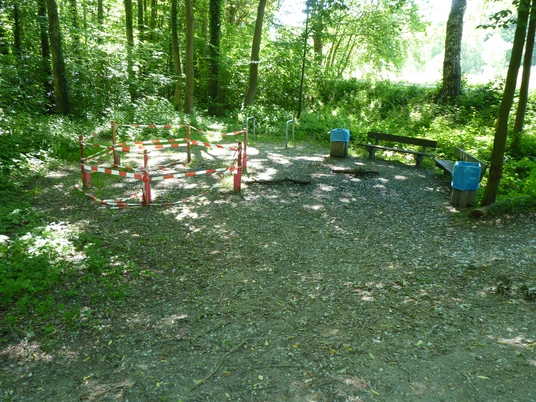 Absperrung um eine beschädigte Fläche im Wald, mit einer angrenzenden Bank und Mülleimern.