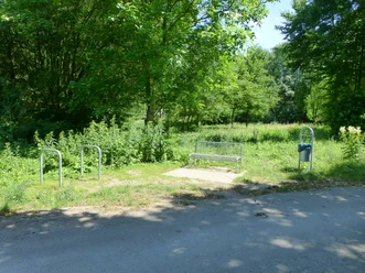 P1120770.JPG Eine Bank auf einem schattigen Platz am Waldrand, umgeben von dichtem Grün und einem Fahrradständer.