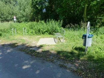 P1120771.JPG Eine kleine gepflasterte Fläche mit einer Metallbank und einem Mülleimer inmitten von Bäumen und Sträuchern.