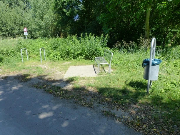 P1120771.JPG Eine kleine gepflasterte Fläche mit einer Metallbank und einem Mülleimer inmitten von Bäumen und Sträuchern.